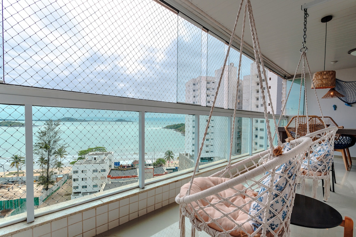 A spacious balcony features comfortable hanging chairs with soft cushions, providing a relaxing space to enjoy coastal views. Large windows allow natural light to fill the area, while the ocean and greenery are visible in the background, enhancing the connection to the outdoors.