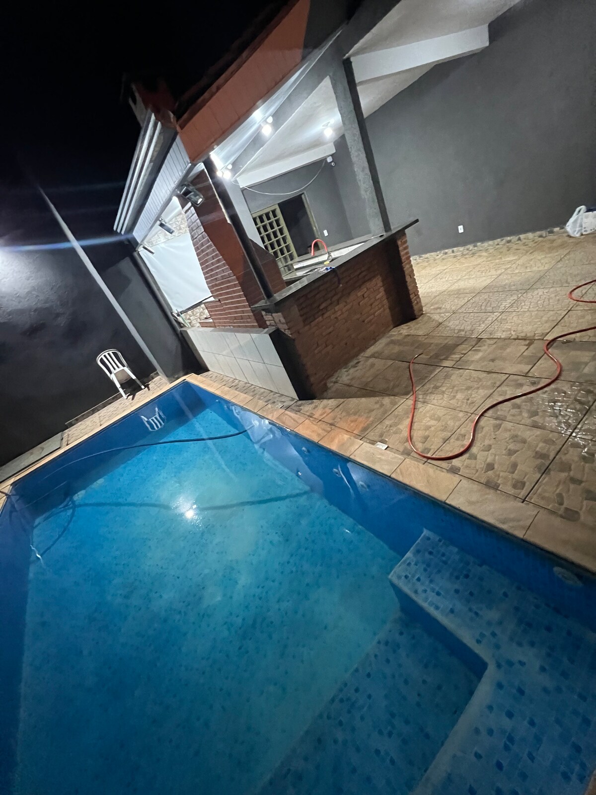 A swimming pool is illuminated at night, showcasing clear blue water. An outdoor kitchen area with a countertop and a white chair is visible nearby, complemented by tiled flooring. Soft lighting accentuates the structural features of the space.