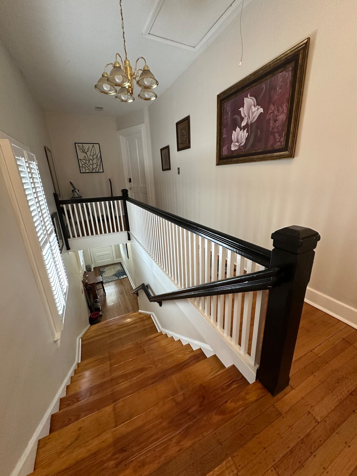 A well-lit staircase features polished wooden steps leading down to the main floor. Black handrails frame both sides, accentuating the white spindles. Light filters in through adjacent windows, illuminating framed artwork on the wall and enhancing the spacious hallway ambiance.