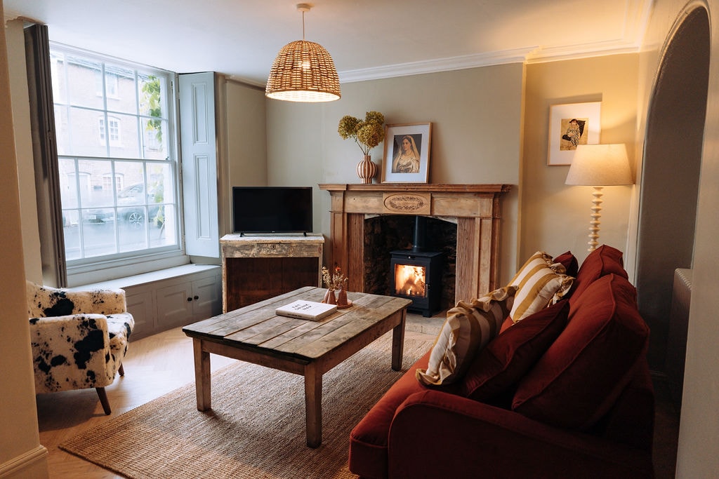A cozy living area features a rustic wooden table positioned in front of a fireplace. A red sofa offers comfortable seating, complemented by a patterned chair. Natural light filters in through a large window, enhancing the warm tones of the decor.