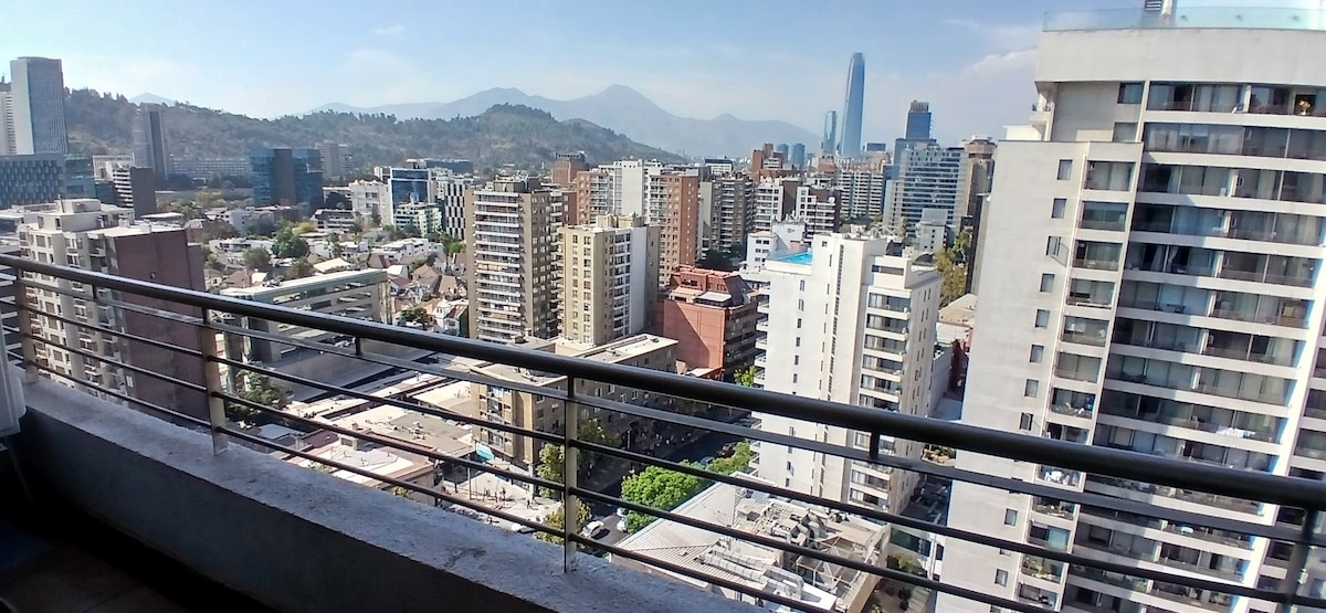 An expansive balcony view captures a vibrant cityscape with a mix of residential and commercial buildings. Surrounding mountains are visible in the background, contrasting with the urban landscape. The railing provides a frame for this elevated perspective of the bustling city below.