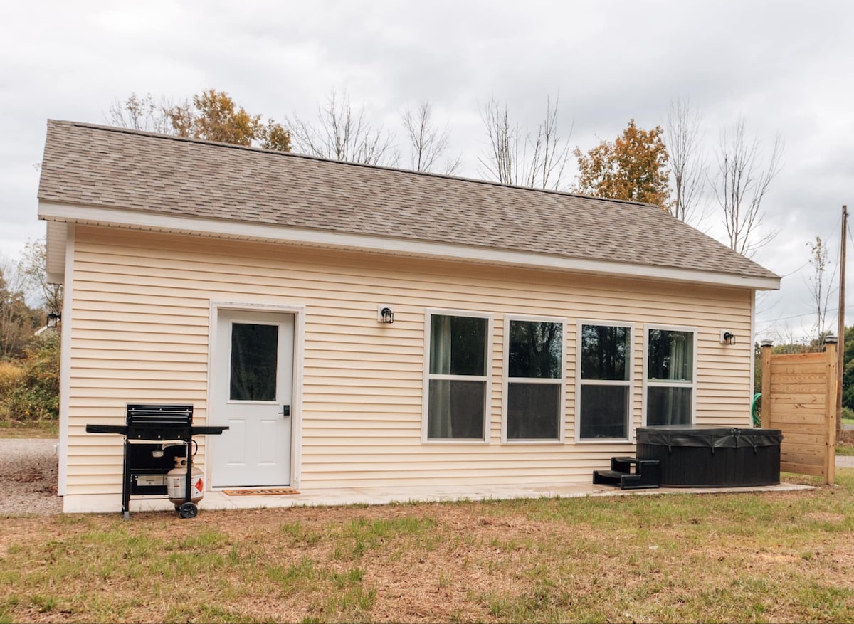 The exterior of a newly constructed tiny home is showcased, featuring a light-colored siding and a sloped roof. Large windows allow natural light to enter, while a grilling area and hot tub are positioned just outside. The surrounding area includes sparse trees and open ground.