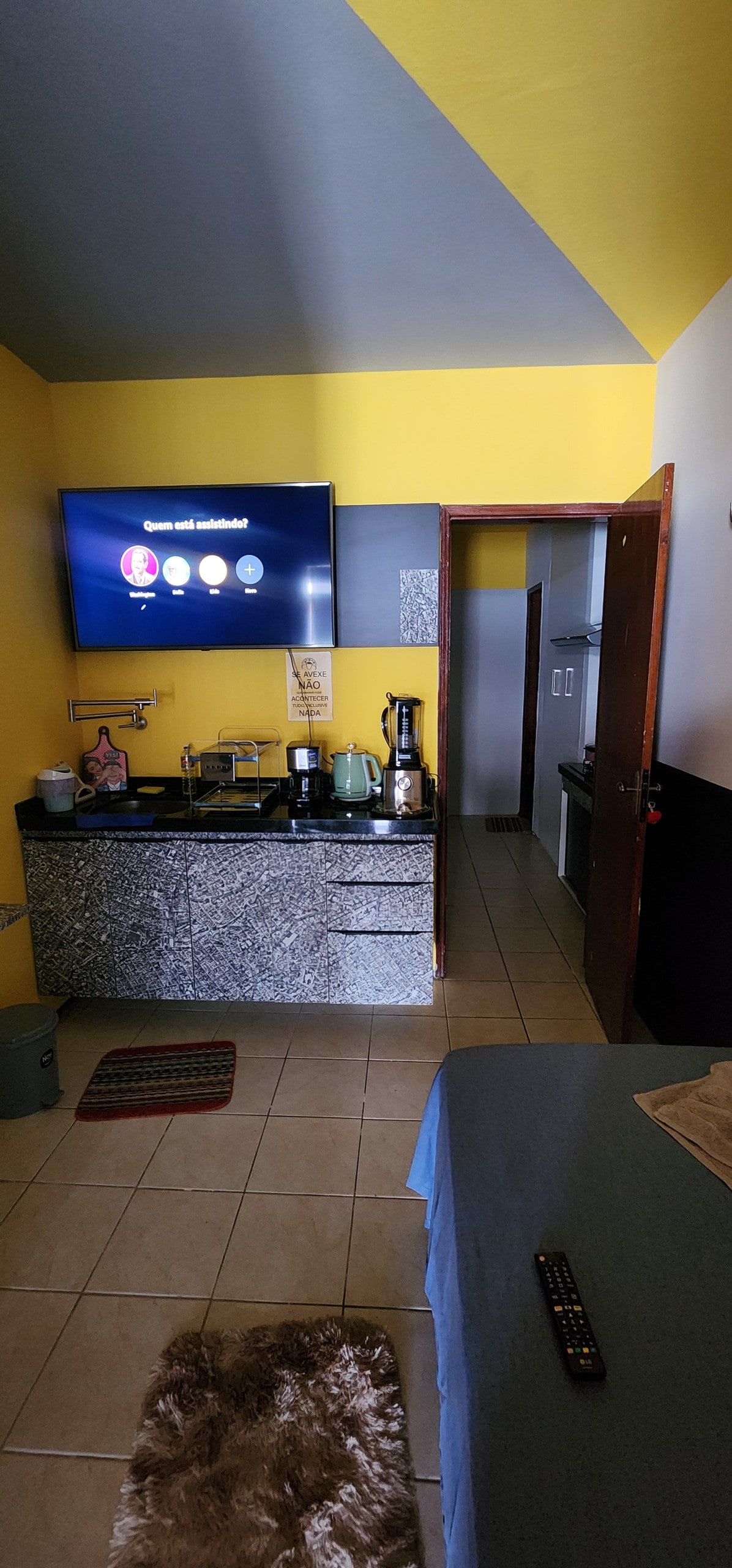A compact living space features a kitchenette with a coffee maker, kettle, and various utensils. A large TV is mounted on the wall, displaying available streaming options. The room is decorated with bright yellow walls, complemented by a cozy bed and soft rug on the tiled floor.