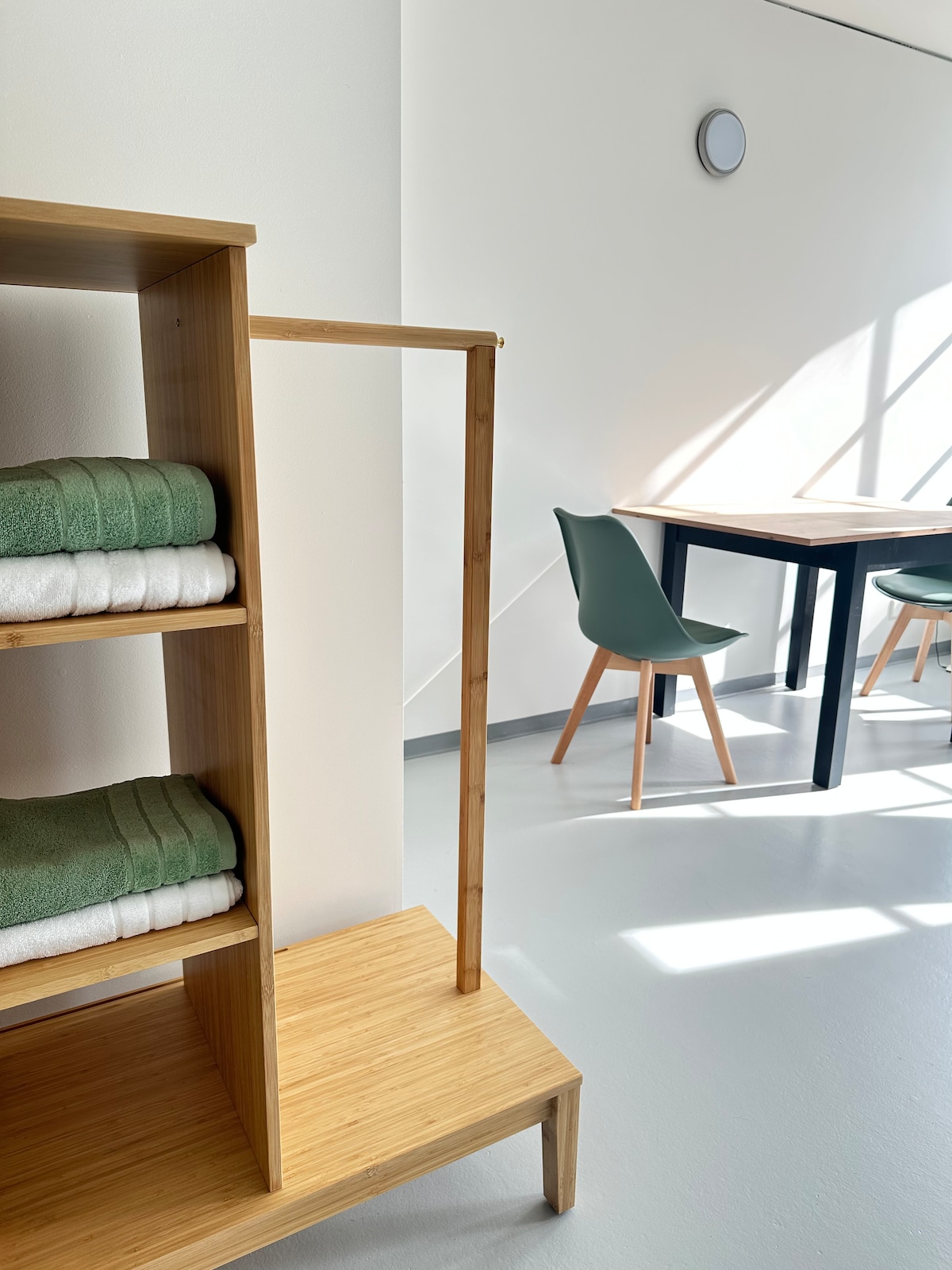 A bamboo shelving unit holds neatly stacked green and white towels. In the background, a dining area features a black table accompanied by two green chairs. Natural light floods the space, illuminating the sleek, minimal design.