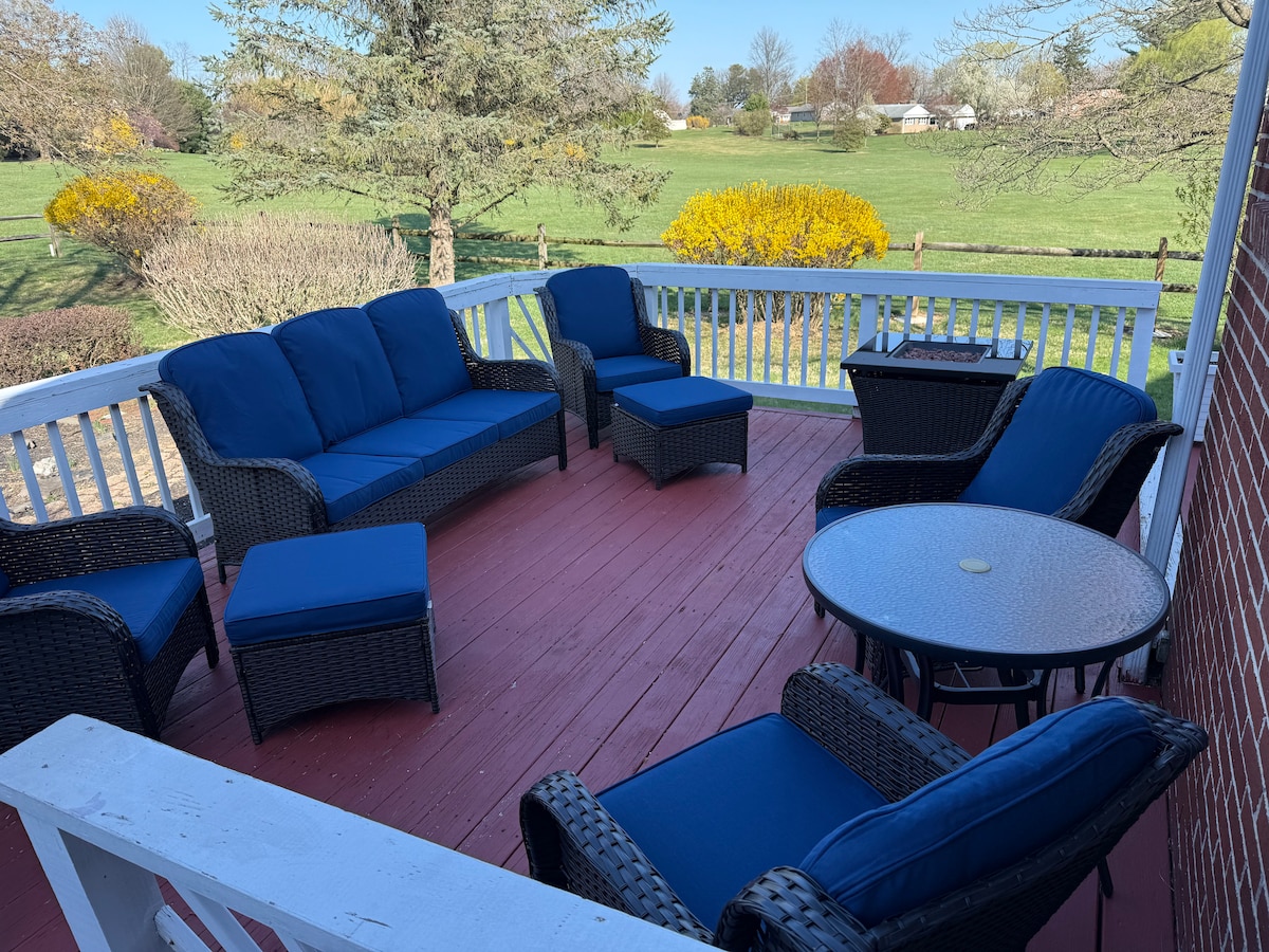 A covered deck area features outdoor seating with navy blue cushions, including a sofa, armchairs, and a coffee table. A scenic view of greenery and flowering bushes can be seen in the background, enhancing the serene outdoor space.