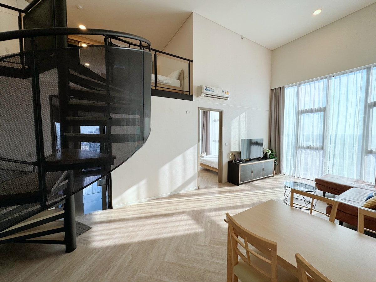 The open-concept living area features large windows allowing natural light to fill the space. A spiral staircase leads to a loft, while a seating area includes a television and a coffee table. A dining table is situated in the foreground, complementing the modern interior design.