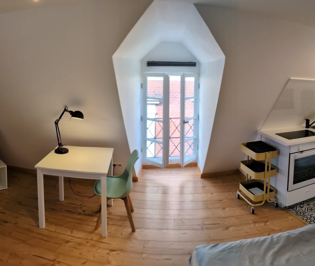 A bright room features a small white table and a soft green chair. A desk lamp is positioned on the table, while a set of double windows with black railings allows natural light to fill the space. The wooden floor adds warmth to the area.