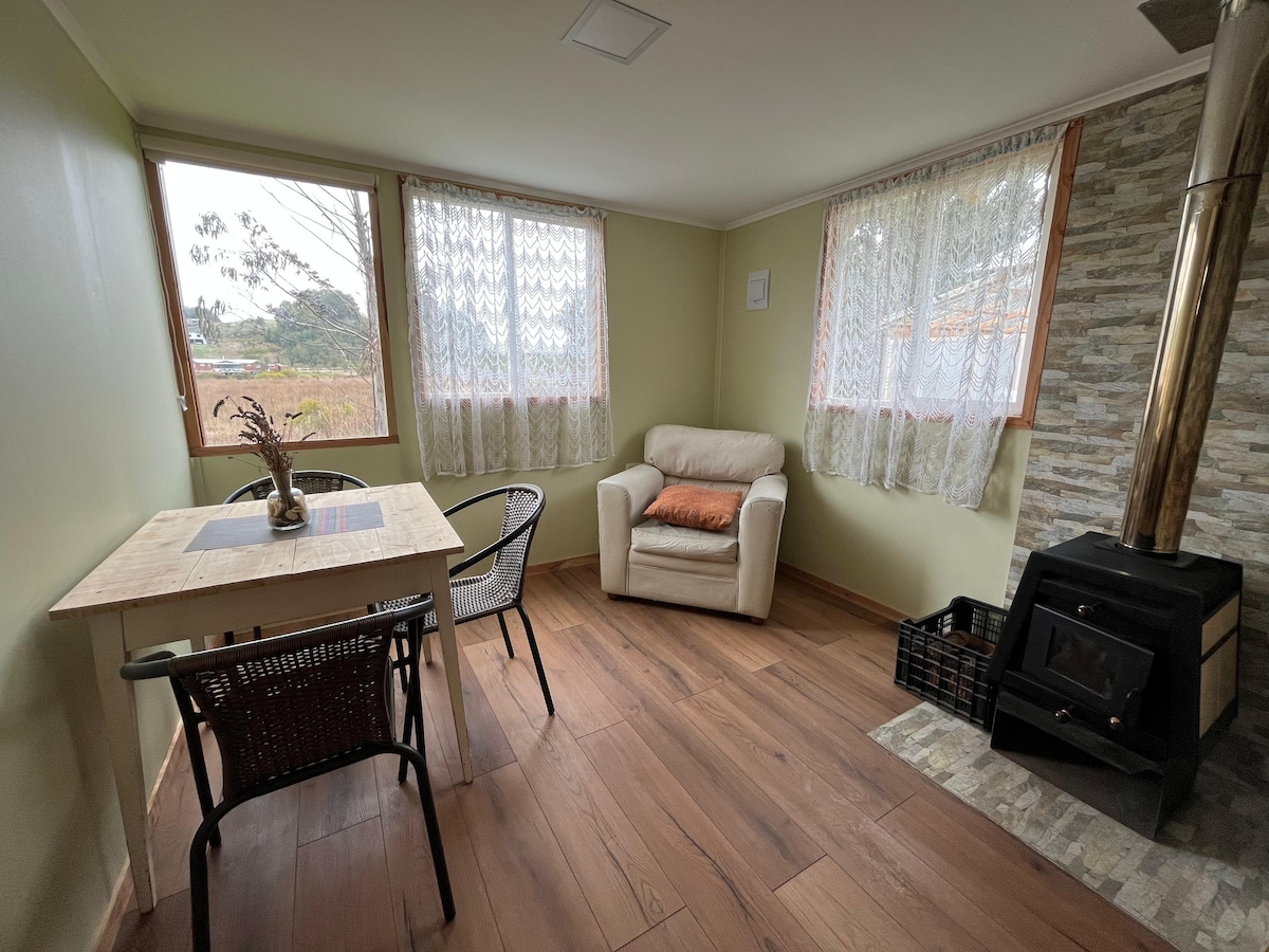 A cozy sitting area features a light-colored armchair positioned near a window with sheer curtains. A small wooden table is accompanied by two black chairs, and a stone fireplace adds warmth to the space. Natural light filters in, enhancing the tranquil atmosphere.
