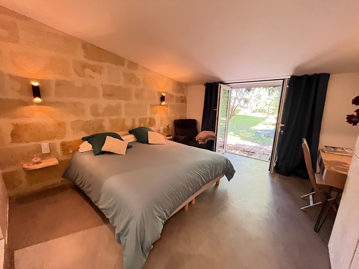 A spacious room is presented with a comfortable double bed covered in soft bedding. Natural light enters through a large glass door leading to a garden view. A cozy armchair is positioned next to the window, complemented by a small desk for workspace.