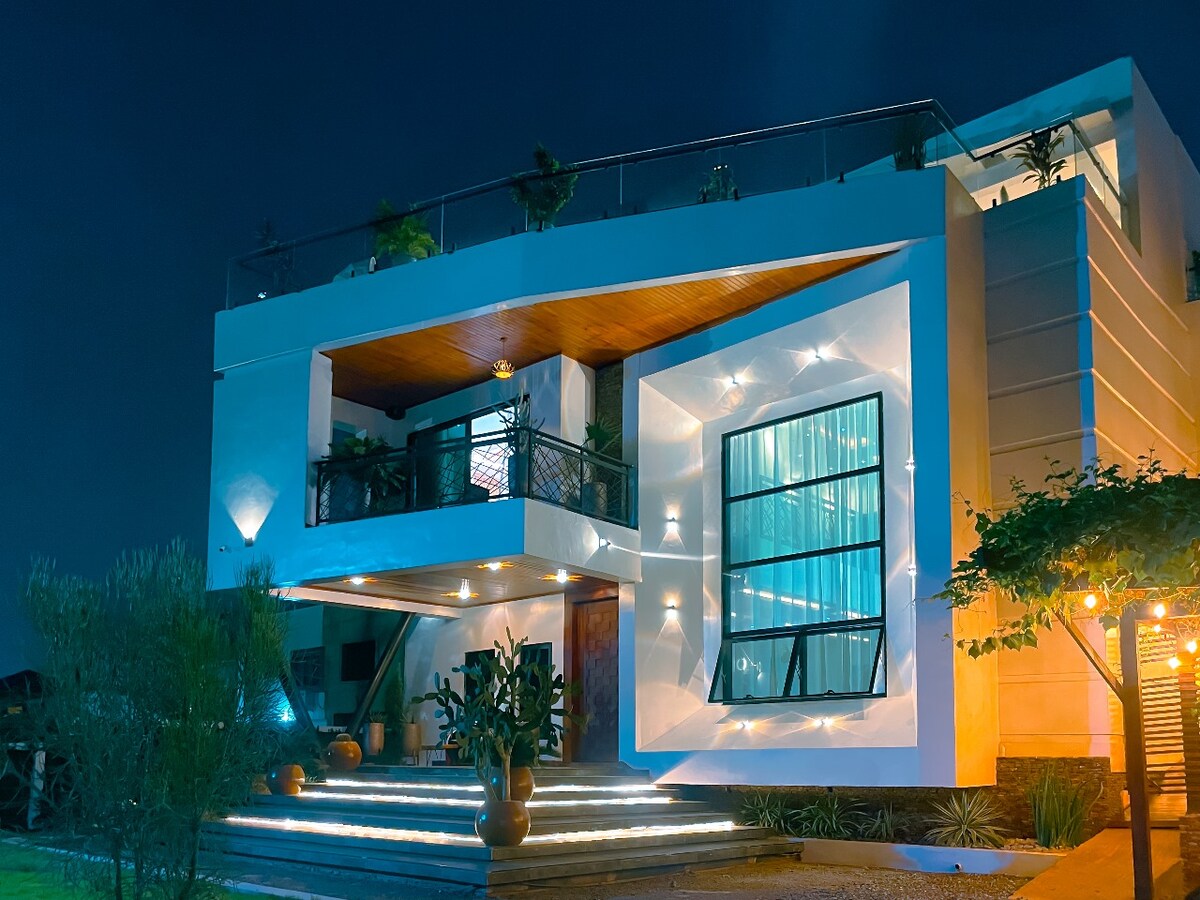 The modern home exterior is illuminated at night, highlighting angular architectural features. Large windows reflect the ambient light, and balconies are adorned with greenery. Softly lit pathways guide guests to the entrance, enhancing the inviting presence of the property.
