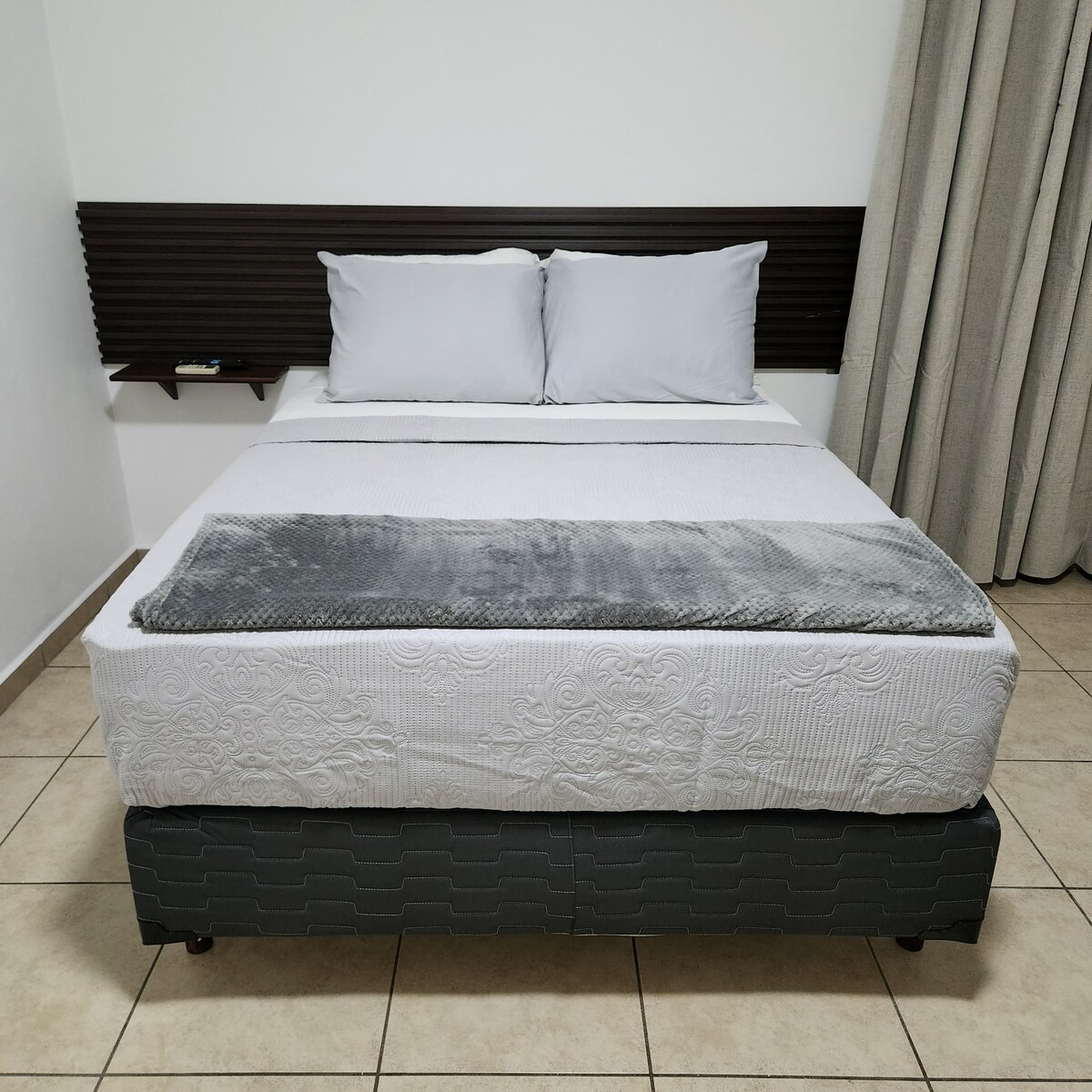 A spacious bed is centered against a plain wall, featuring a minimalistic headboard. Light grey pillows rest against the headboard, and a soft throw blanket is neatly folded at the foot of the bed. Neutral-colored curtains frame the window beside it.