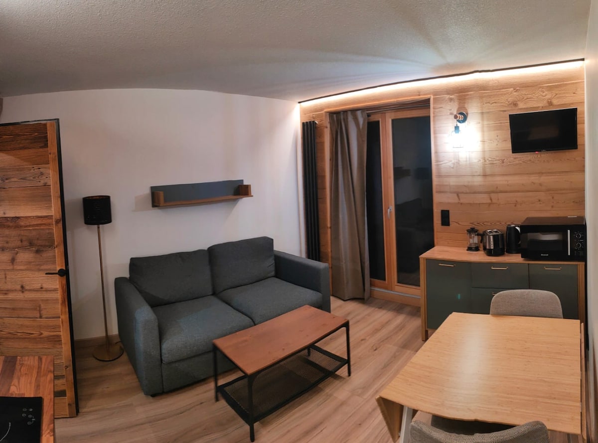 A modern living area is furnished with a gray sofa and a wooden coffee table. Natural light enters through a large window, adorned with curtains. A kitchenette is equipped with essential appliances, including a microwave and coffee maker, complemented by a dining table and chairs.