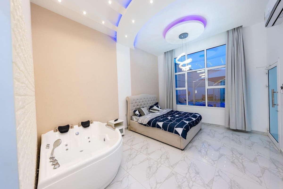 A spacious room features a comfortable bed with a patterned duvet and a stylish headboard. A modern jacuzzi adds to the relaxation options. Large windows allow natural light to fill the space, with soft curtains partially drawn, and recessed lighting enhances the calming ambiance.