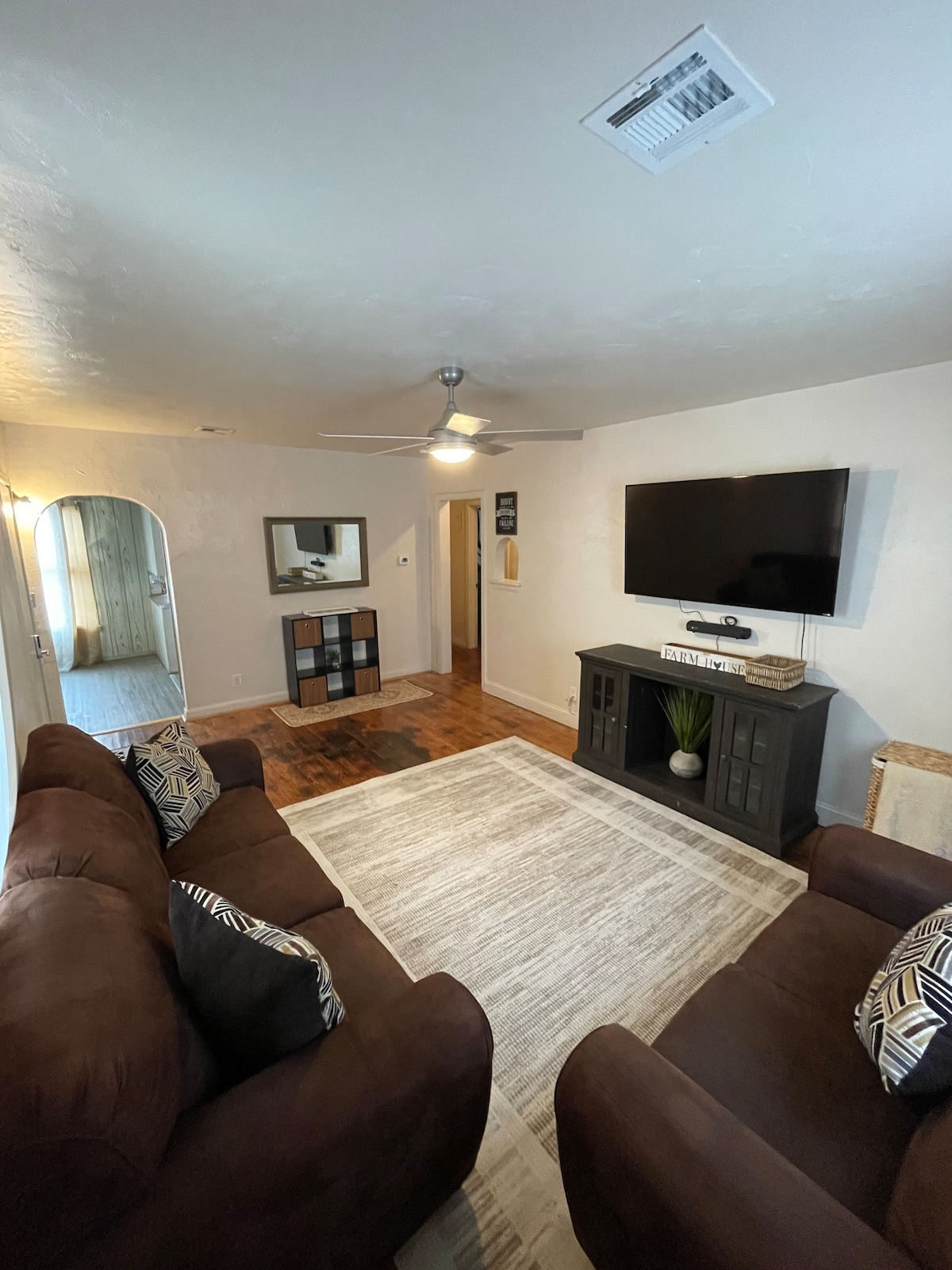 The living area features a comfortable brown sofa with decorative cushions and a light area rug. A flat-screen TV is mounted on the wall, above a dark wooden entertainment unit. Natural light comes in from a nearby window, complementing the warm, inviting space.