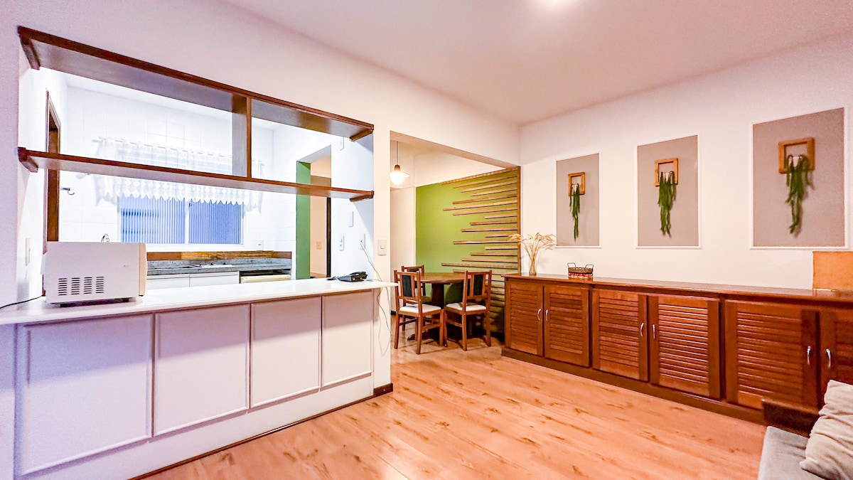 A spacious living area showcases a contemporary open kitchen with a microwave and clear counters. Dining space features a wooden table surrounded by chairs. Natural wood storage units provide ample space, while unique wall decor offers a touch of greenery to the inviting atmosphere.