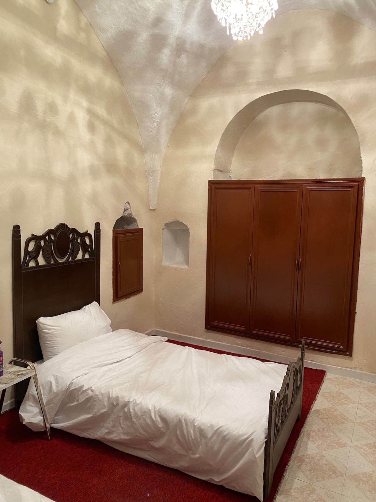 A charming sleeping area features an arched ceiling with soft, natural light filtering in. A spacious bed is positioned against a wall, complemented by a wooden headboard. A wooden wardrobe occupies one section, while decorative alcoves add interest to the cream-colored walls.