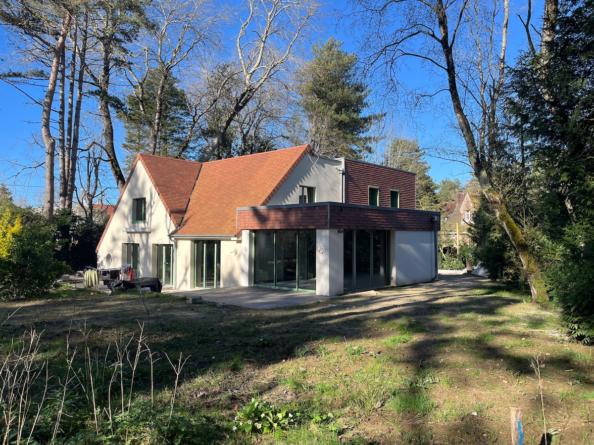 The exterior of a modern house is presented, featuring a combination of sloped and flat roofs. Large glass windows allow natural light into the living spaces. The surrounding area includes green landscaping with trees, offering a sense of privacy and tranquility.
