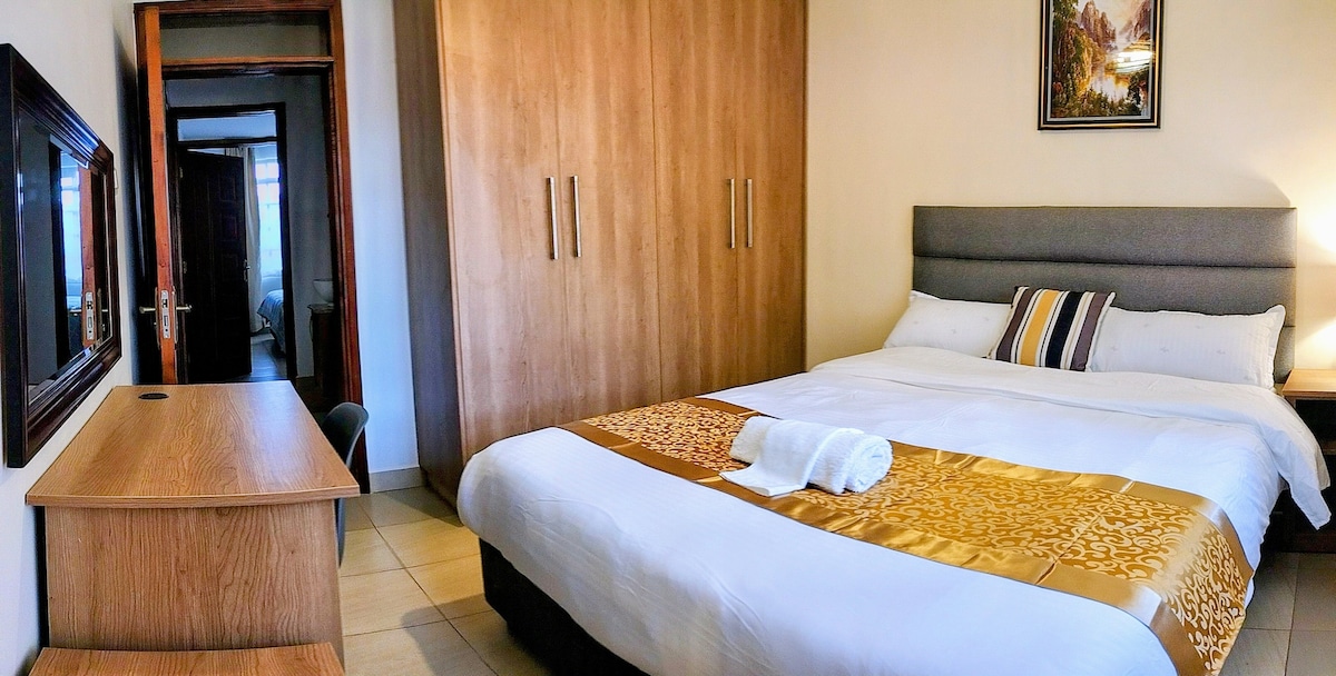 A spacious sleeping area features a large bed dressed in crisp white linens, complemented by an elegant golden throw. A decorative painting hangs above the headboard, while a wooden desk and mirror add functionality to the room.