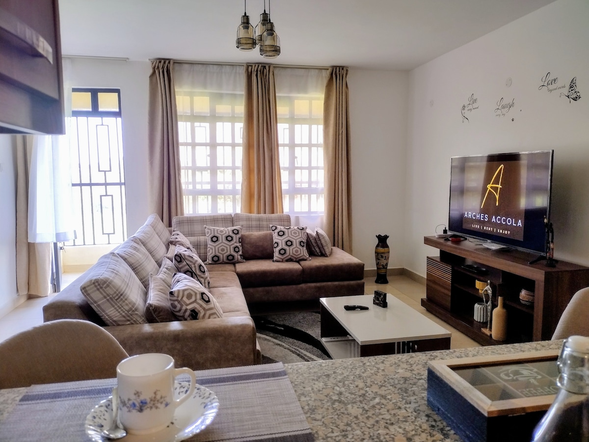 A comfortable living area features a sectional sofa adorned with patterned cushions. A flat-screen TV is mounted on a wooden console beneath decorative accents. Natural light flows through the large windows, which are framed by sheer and solid curtains.