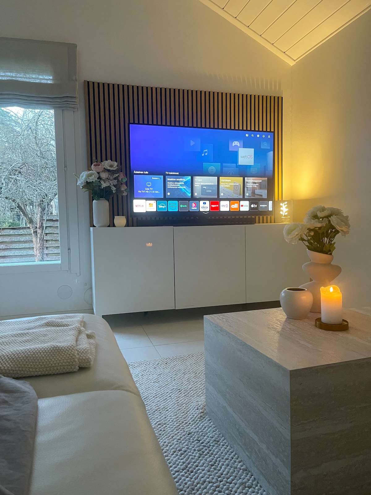 A modern living area features a sleek television mounted on a wooden panel. Soft lighting creates a calming ambiance, complemented by decorative flowers and two lit candles on a stone coffee table. A plush throw blanket is casually placed on the nearby sofa.