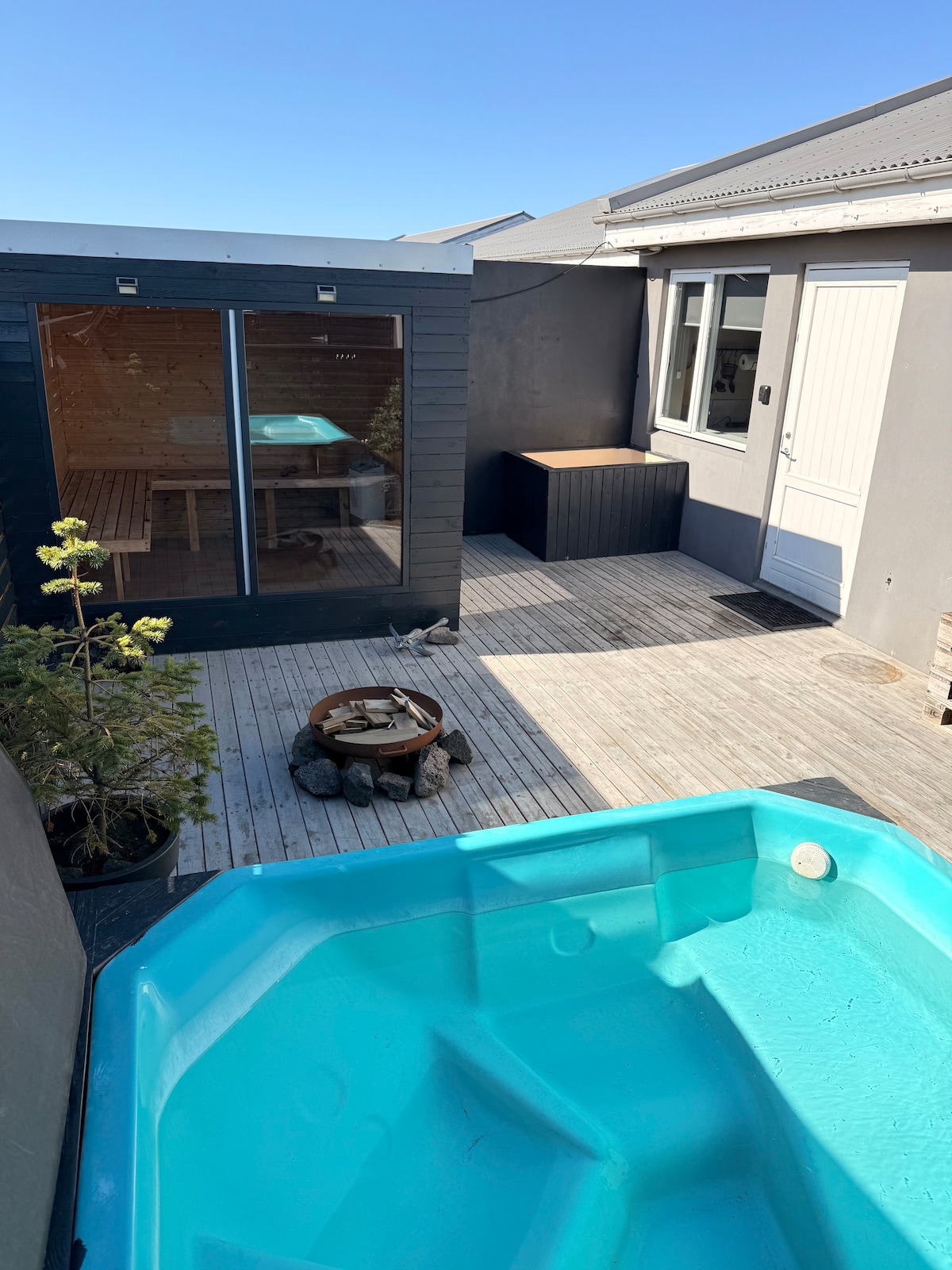 A private spa garden is presented, featuring a hot tub in turquoise waters and a wooden patio. A sauna structure is visible in the background, alongside a small fire pit surrounded by stone. Natural light illuminates the area, emphasizing the serene atmosphere.