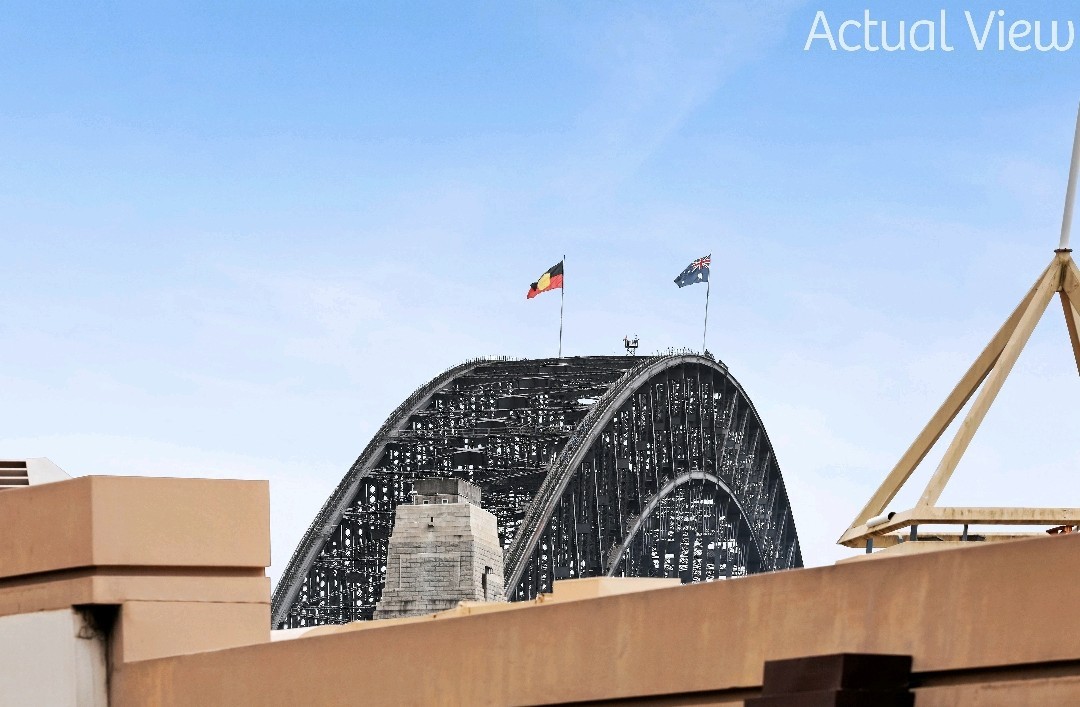 The image showcases a view of the Harbour Bridge, with its distinctive arch prominently displayed. Flags are visible at the top, representing Germany and Australia. The foreground includes part of the terrace, blending with the urban landscape.