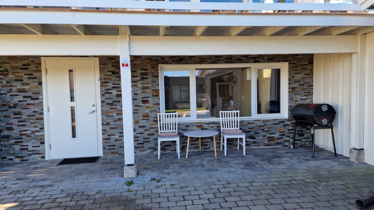 An outdoor seating area features a round table accompanied by two white chairs, set against a stone wall. A charcoal grill is positioned nearby, providing an option for outdoor cooking. The entrance to the unit is visible, framed by a porch roof.