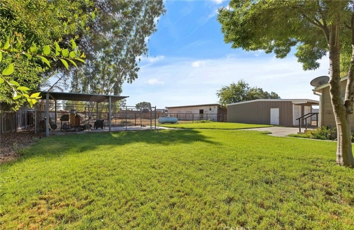 A spacious yard is presented with green grass and several trees for shade. Two covered areas are visible, alongside a large parking area. Fencing outlines the property, and additional structures are seen in the background, adding to the outdoor functionality of the space.