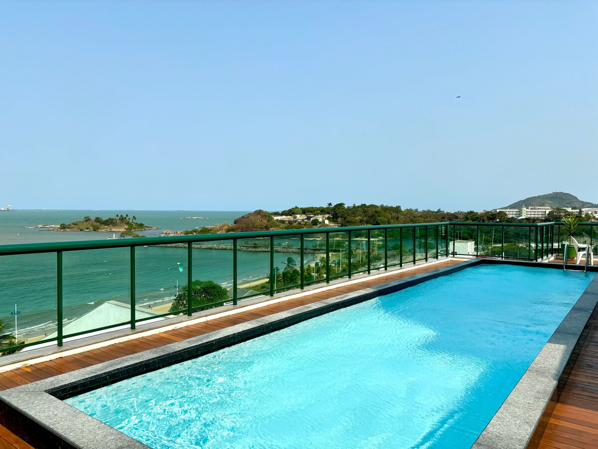 A rooftop area features a swimming pool surrounded by a glass railing, providing views of the ocean and coastal landscape. The tranquil water reflects the sky, inviting relaxation in a serene environment.