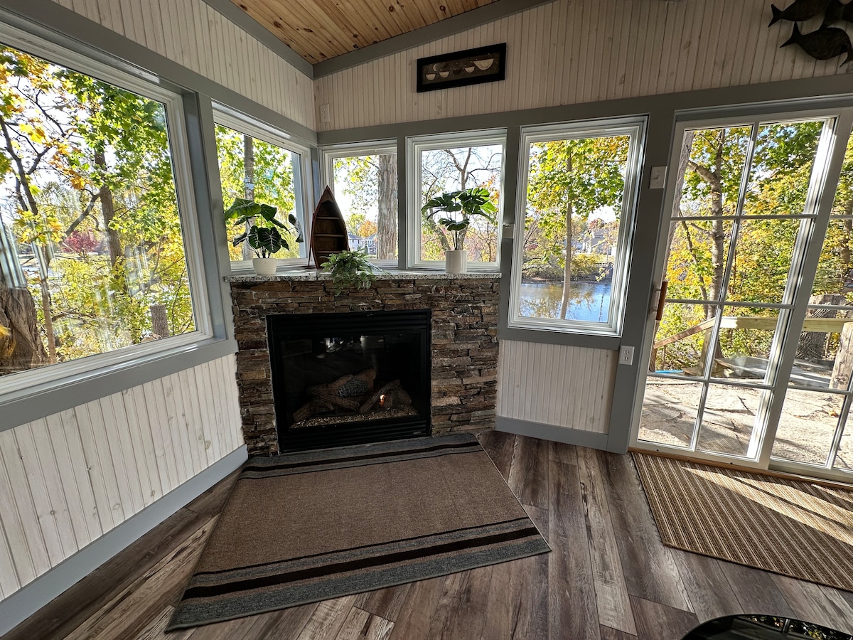 A sunroom is highlighted by large windows that provide views of the surrounding nature and river. A stone fireplace adds warmth to the space, complemented by a beige area rug on the wooden floor. Potted plants decorate the windowsills, enhancing the inviting atmosphere.
