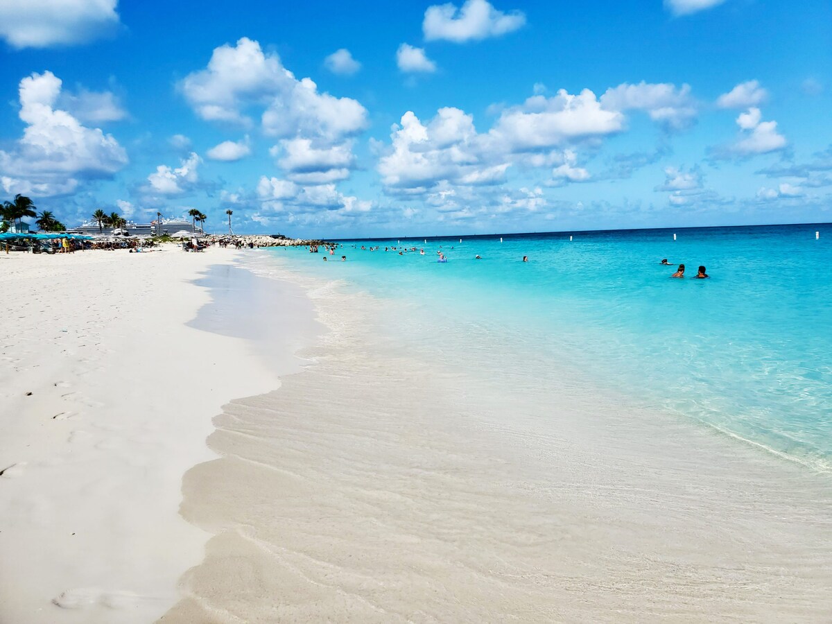 A wide stretch of sandy beach features soft white sand meeting clear turquoise waters. Gentle waves roll ashore under a bright blue sky filled with fluffy white clouds. People can be seen enjoying the water, creating a lively yet serene atmosphere.