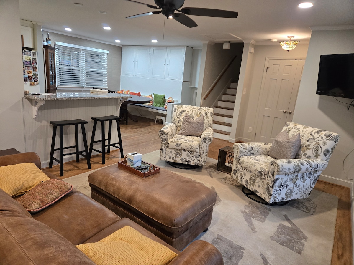 A shared living space contains a comfortable leather sofa and two patterned chairs. A coffee table sits in the center, surrounded by a rug. A bar with two stools is visible, and an area in the background showcases a staircase and large windows with natural light.