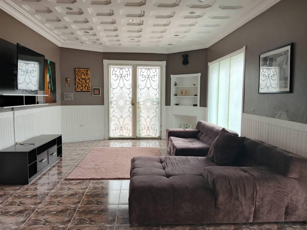 The living room features a plush sectional sofa positioned for comfort, complemented by a large area rug beneath. Natural light filters in through the glass doors, which open to a balcony with decorative wrought iron accents. A television unit and shelving are also present.