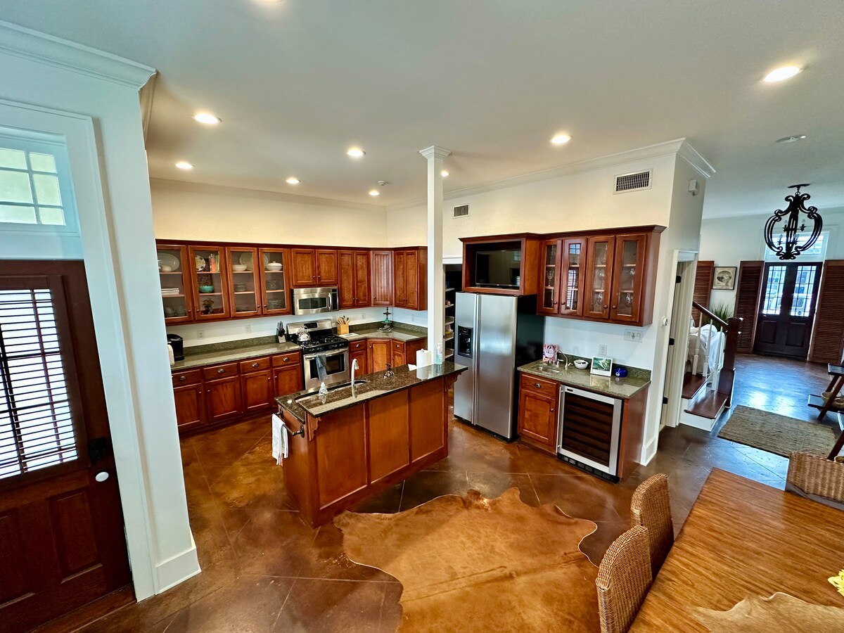 A modern kitchen features rich wooden cabinetry and stainless steel appliances. A large island with a granite countertop stands at the center. Natural light fills the space, enhancing the clean lines and functionality of the design. A dining area with woven seating is visible in the background.
