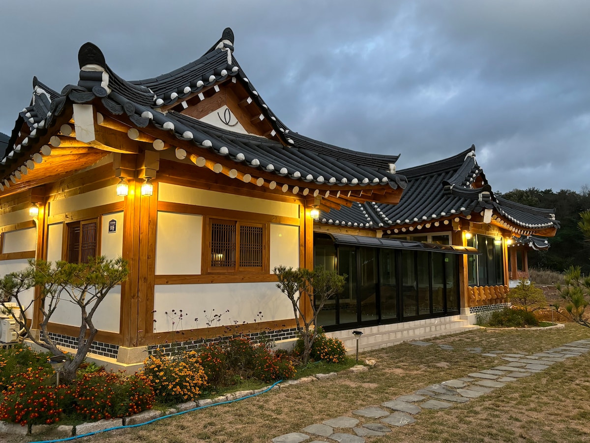 A traditional Hanok structure is depicted, showcasing distinct curved roofs and wooden beams. Soft exterior lighting highlights the intricate architectural details. Well-maintained flower beds and a stone pathway frame the house, creating a harmonious connection with the natural surroundings.