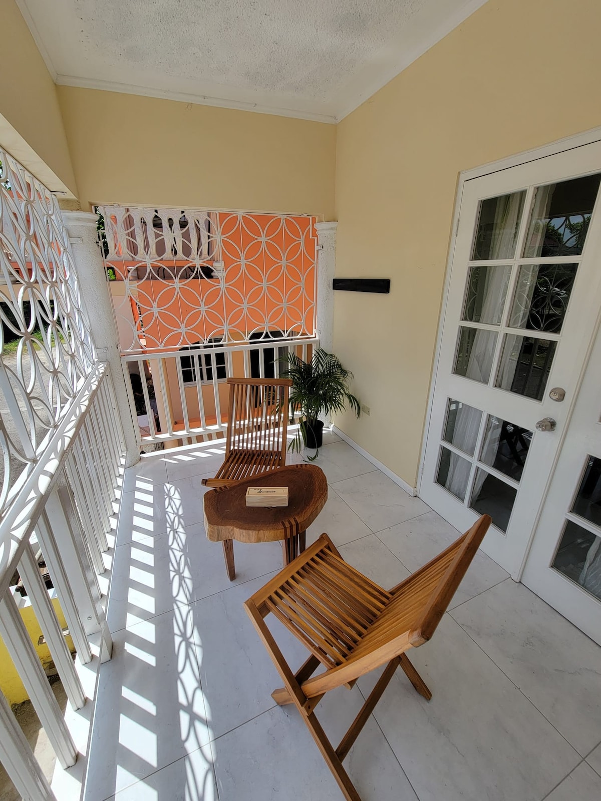 A small balcony area is presented, featuring two wooden chairs and a round table. Natural light filters through a patterned screen, casting gentle shadows on the tiled floor. A potted plant adds a touch of greenery to the space, enhancing the overall atmosphere.