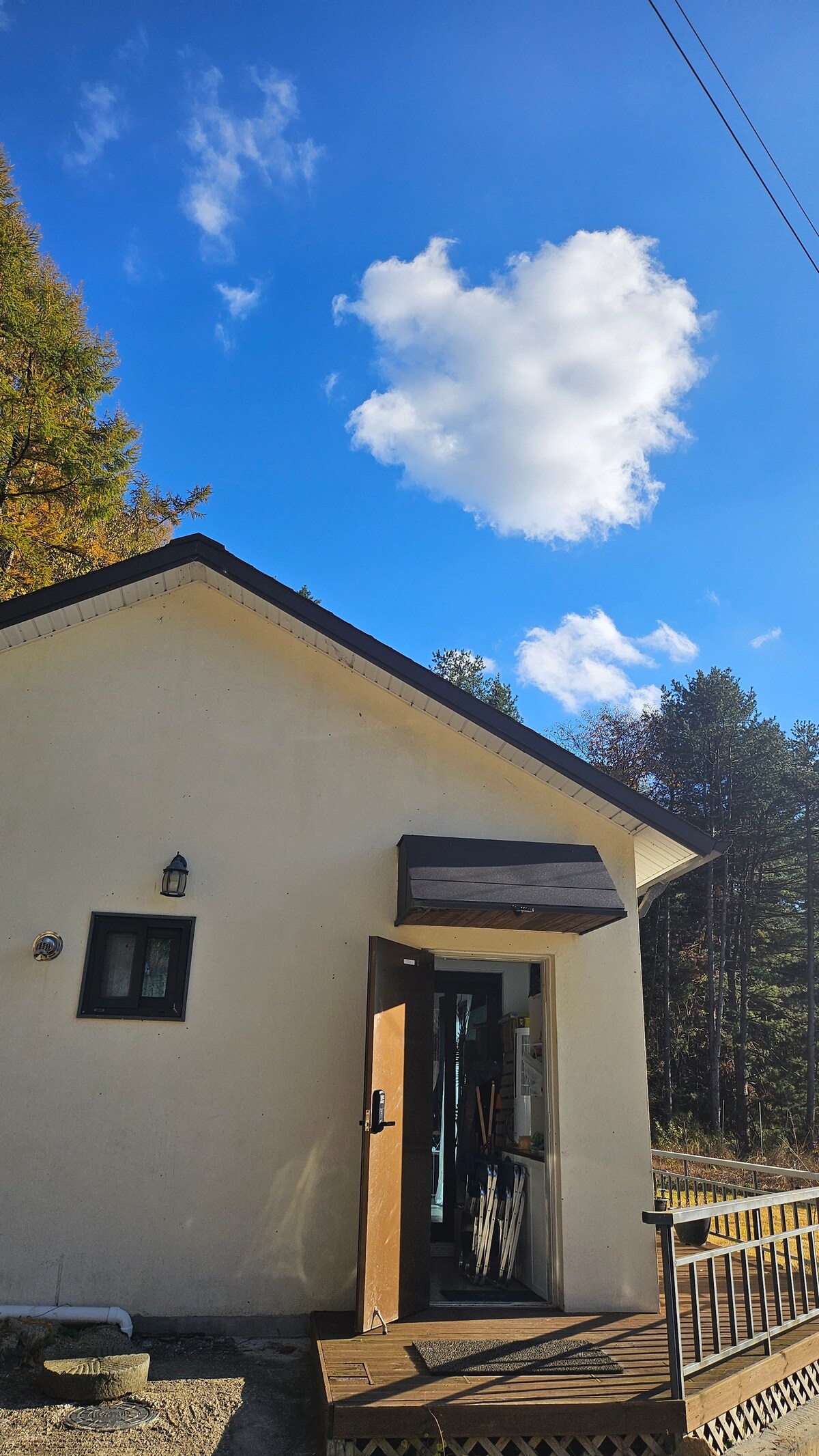 The exterior of a cozy house is shown, framed by a clear blue sky and scattered trees. A wooden deck leads to an inviting front door, which is slightly ajar, allowing a glimpse inside. A single cloud floats above, adding to the tranquil setting.