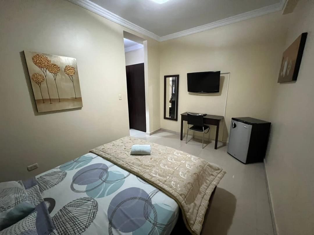 A well-organized bedroom features a spacious bed with decorative bedding. A wall-mounted television and a small desk are present, while a compact refrigerator is positioned nearby. Natural light enters through a doorway, highlighting the neutral wall colors and artwork.