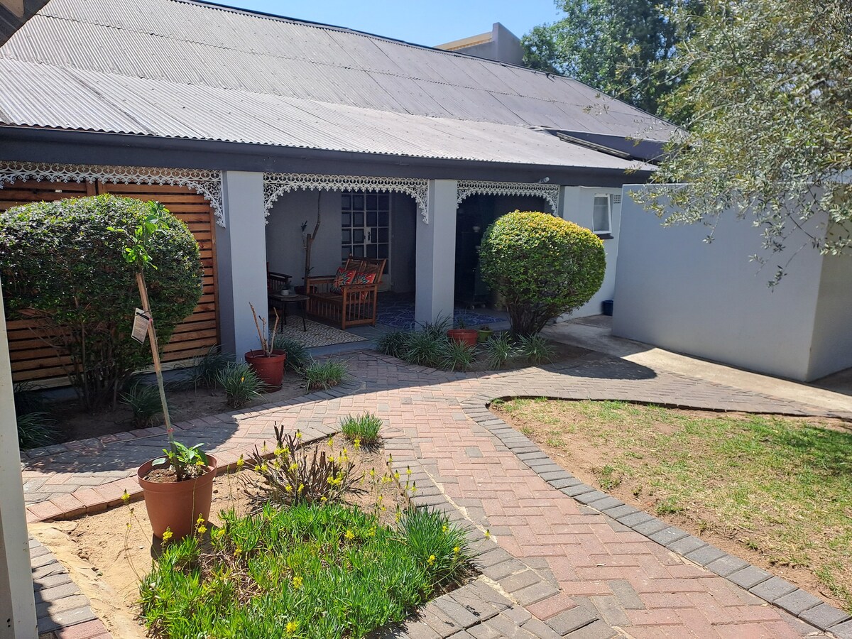 A charming exterior pathway leads to a covered porch, framed by neat shrubs and pots with plants. The entrance is flanked by two inviting rock chairs, offering a cozy spot for relaxation. The overall setting is surrounded by greenery, enhancing the peaceful ambiance.