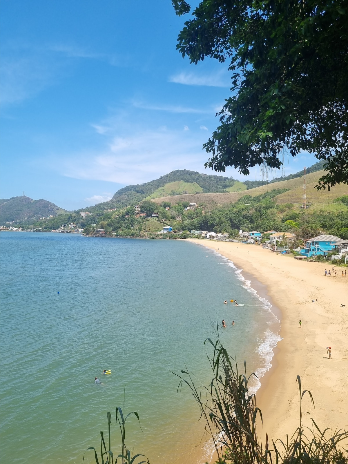 A serene beach scene is displayed with golden sand gently meeting the turquoise waters. Lush green hills rise in the background, and a few colorful structures are visible along the shore. Small figures can be seen enjoying the water and sand under a clear blue sky.