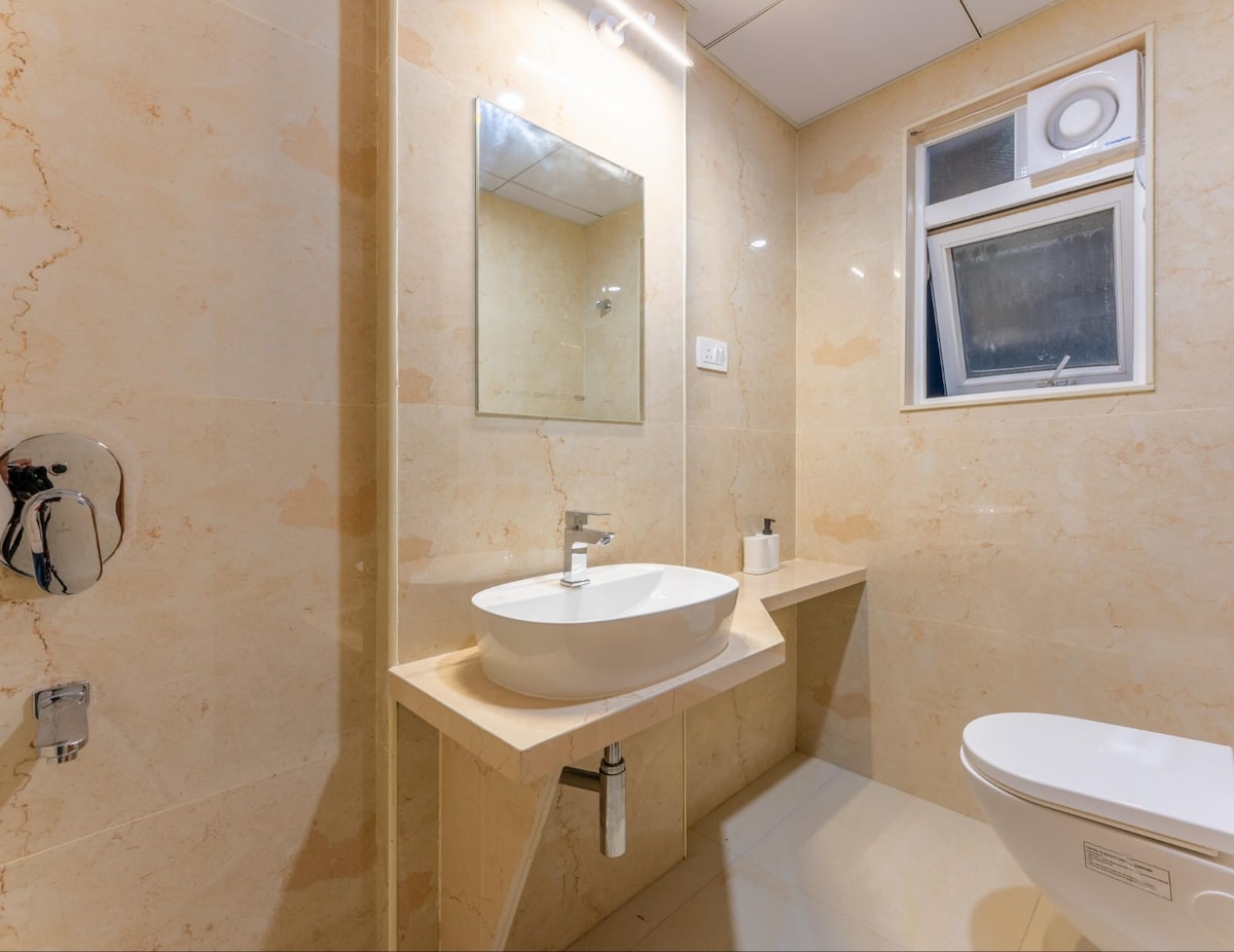 A modern bathroom features beige stone walls and a sleek sink with a mirror above it. Natural light enters through a small window, while a minimalist toilet and built-in shelf for toiletries is visible. Fixtures are neatly arranged, emphasizing the space's functionality.