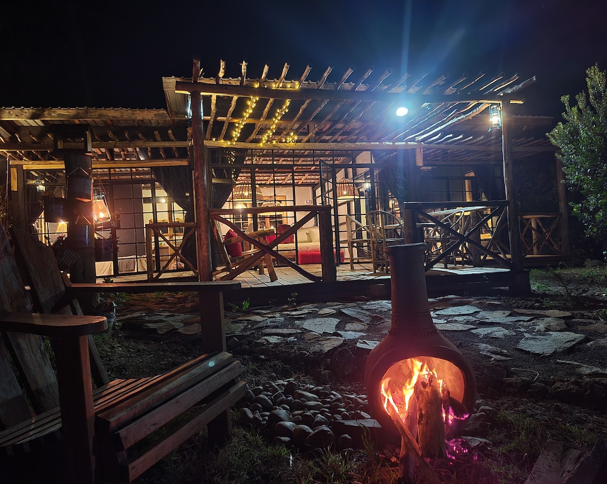 The cabin is illuminated softly at night, with a fire burning in a chiminea in the foreground. A wooden bench is positioned nearby, creating a cozy setting. The warm glow highlights the wooden structure, showcasing an inviting entry with open space leading to the interior.