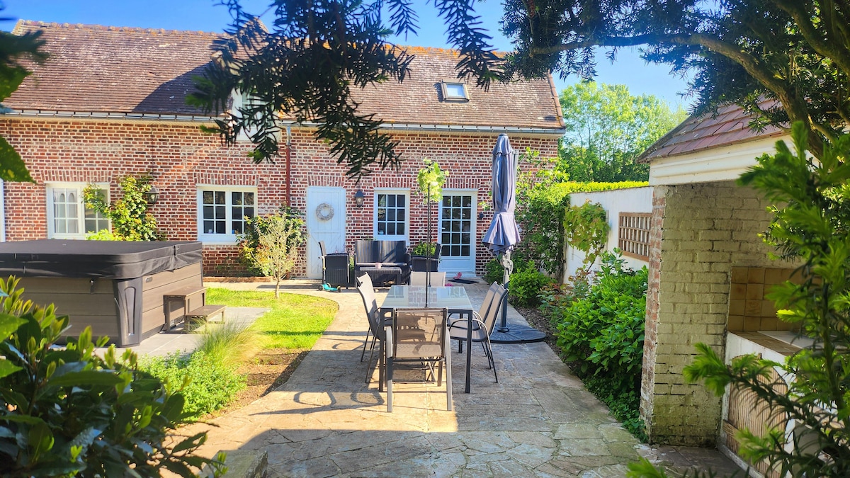 A charming outdoor space is featured, showcasing a patio with a dining table and chairs surrounded by greenery. A hot tub is visible to the left, complemented by a sun umbrella. The brick facade of the building is partially obscured by trees.