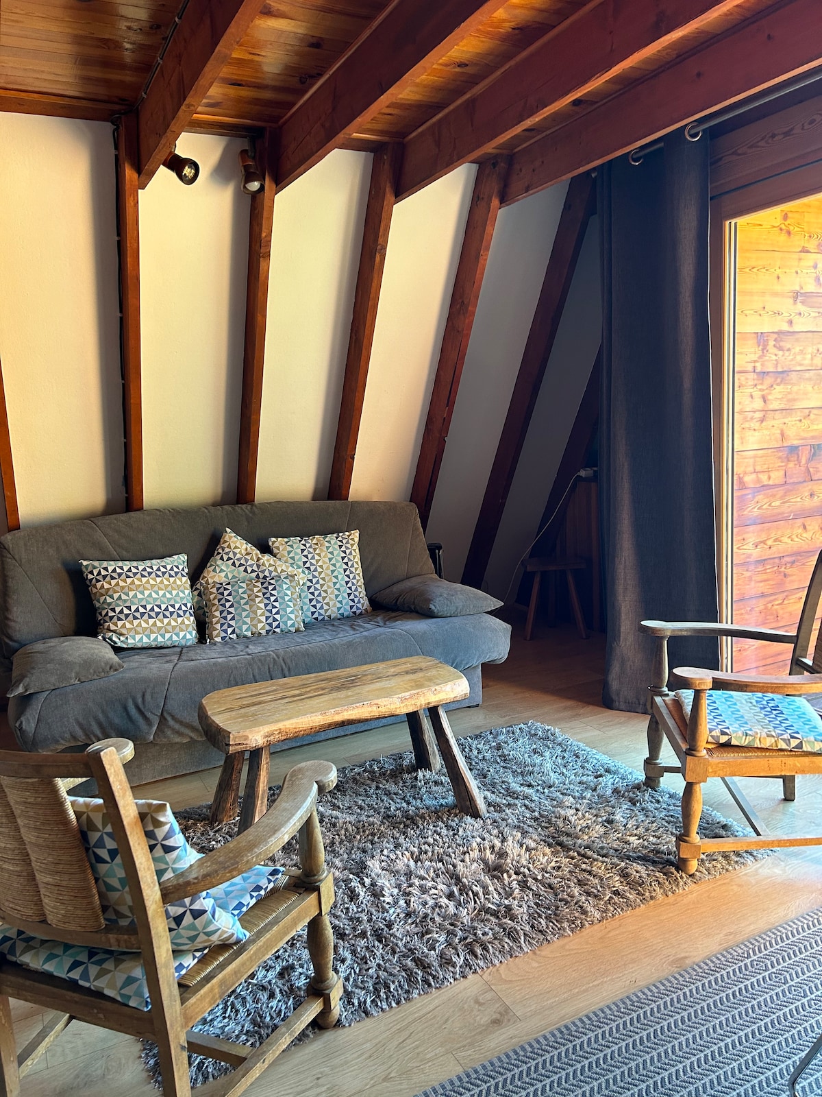 A cozy living area is presented, featuring a grey sofa adorned with patterned cushions. A small wooden table occupies the center, surrounded by two armchairs. Natural light is allowed in through the large doors, highlighting the wood-beamed ceiling and soft area rug beneath the furniture.