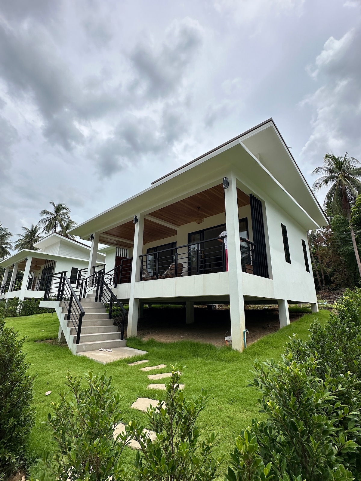 A modern bungalow with a welcoming porch is surrounded by lush greenery. Steps lead up to the elevated entrance, while palm trees provide a natural backdrop. The flat roofline and large windows enhance the airy feel of the home.