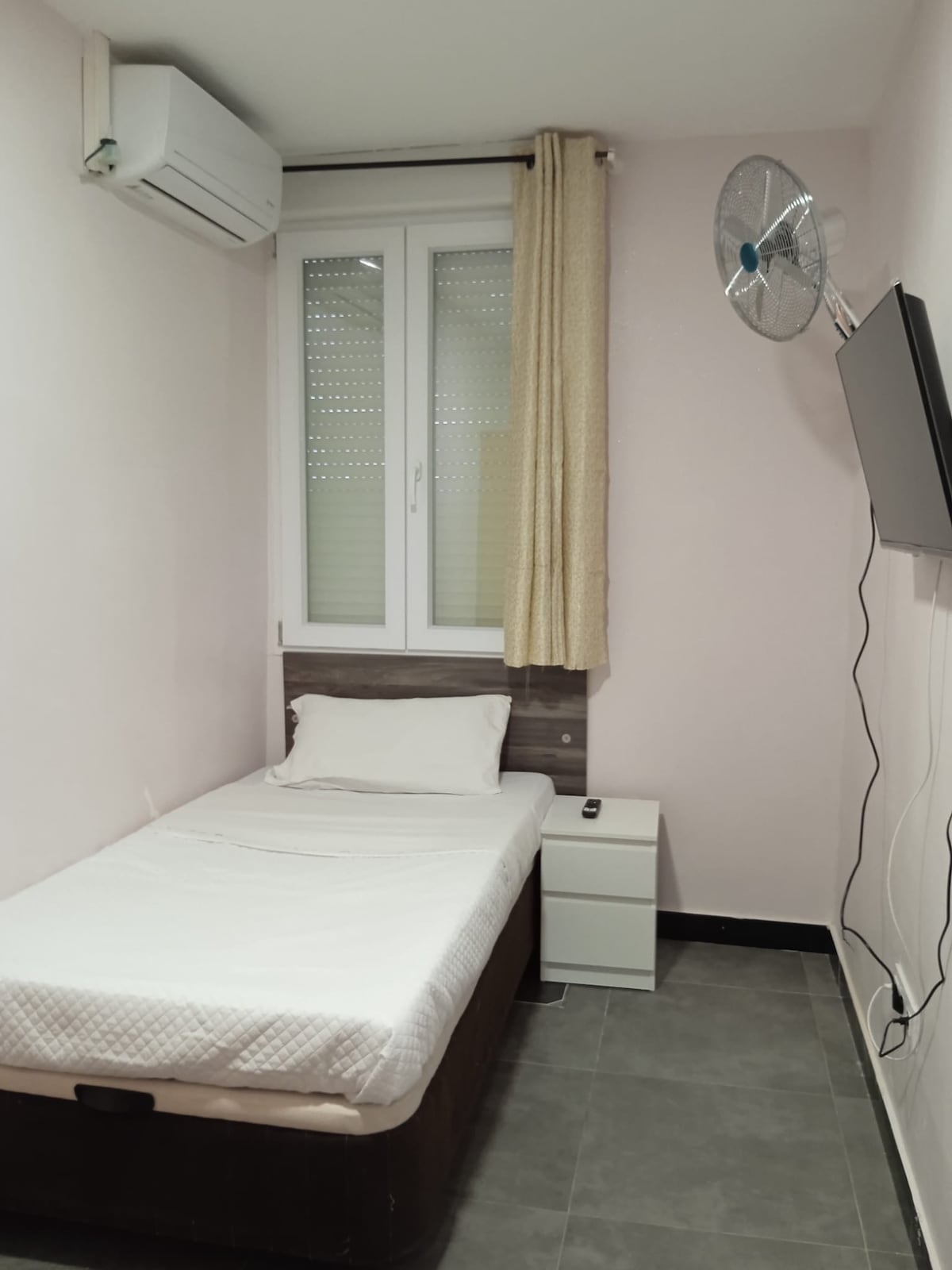 A compact guest room features a single bed with a neutral-toned mattress. A white nightstand is positioned beside the bed. A flat-screen television is mounted on the wall opposite the bed, and a ceiling fan provides additional air circulation. Natural light enters through a window with sheer curtains.