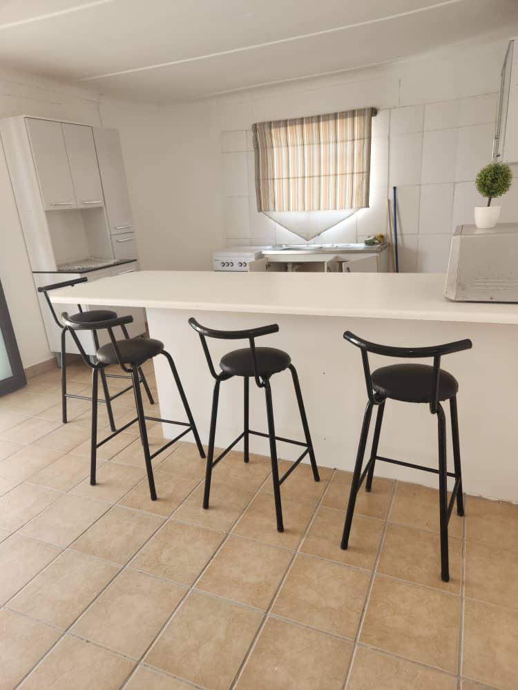 A bright kitchen space is highlighted by a central island bar with four high stools. Natural light filters through a window adorned with a patterned curtain, and cabinetry is arranged along one wall, contributing to a functional layout.
