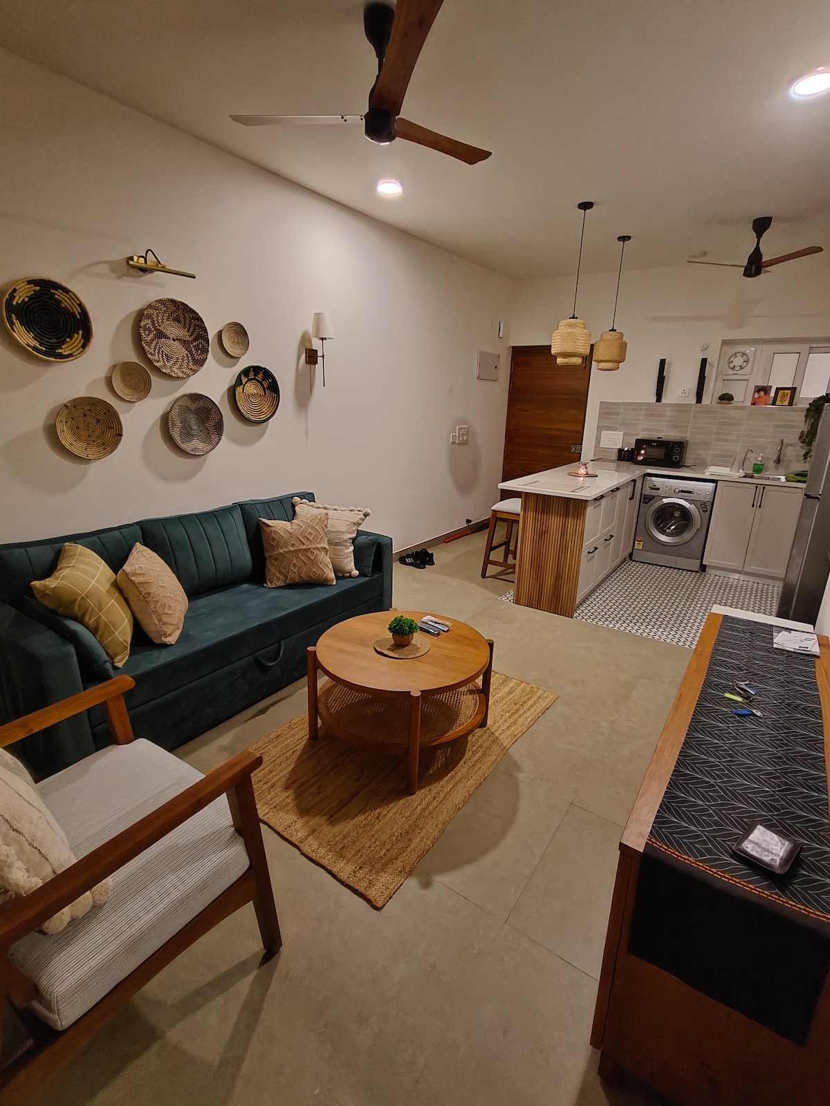 A living area showcases a green sofa adorned with patterned cushions, alongside a wooden chair. A round coffee table rests on a woven mat. Decorative plates are hung on the wall, and the kitchenette is visible in the background with a washing machine and modern fixtures.