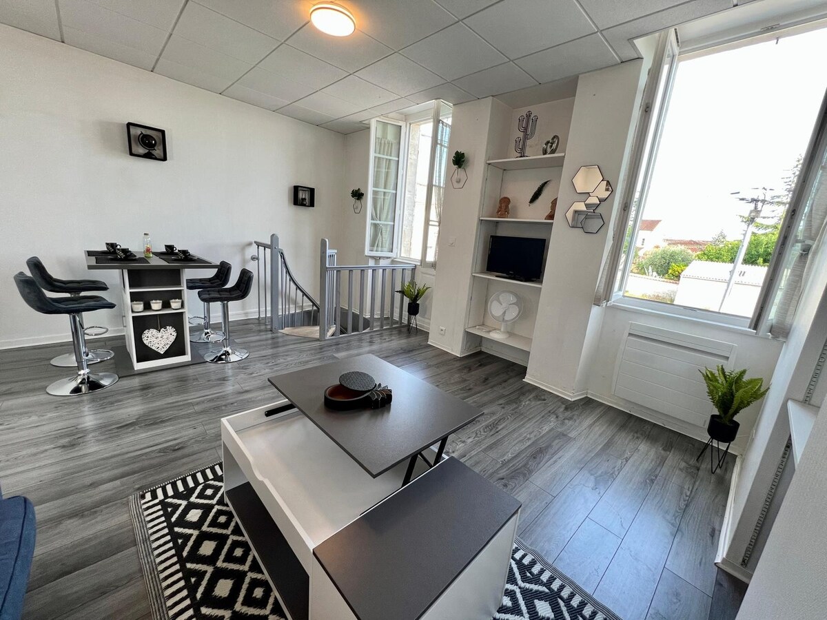 The living area features a modern layout with a cozy seating arrangement and a sleek coffee table. Natural light filters through large windows, illuminating the space. A dining bar with black stools is positioned nearby, offering a functional and stylish area for meals.