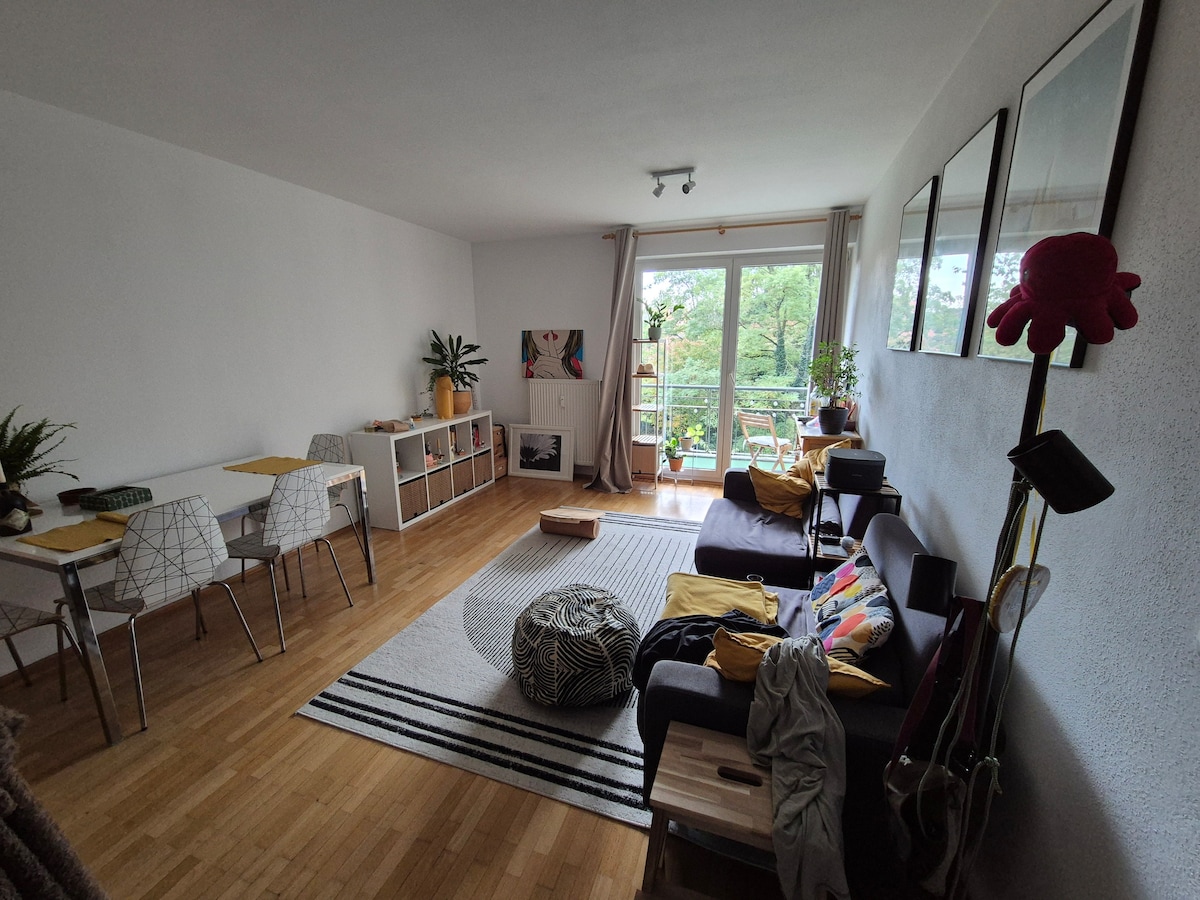 A bright living area features a comfortable sofa adorned with colorful cushions and a plush rug. A dining table with chairs is positioned nearby, while large glass doors open to a greenery-filled balcony. Shelves display decorative items and plants, enhancing the cozy ambiance.